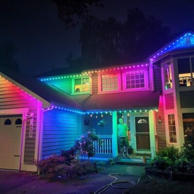 A residential home at night featuring multicolored LED lights along its exterior and a person on the front porch