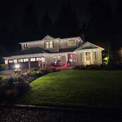 Exterior view of a two-story house at night with permanent colorful lights installed along the roof and festive decorations on windows.