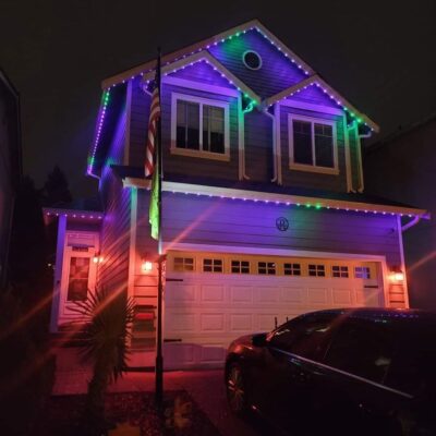 A two-story house at night lit by permanent multicolored holiday lights with an American flag and a parked car in the driveway