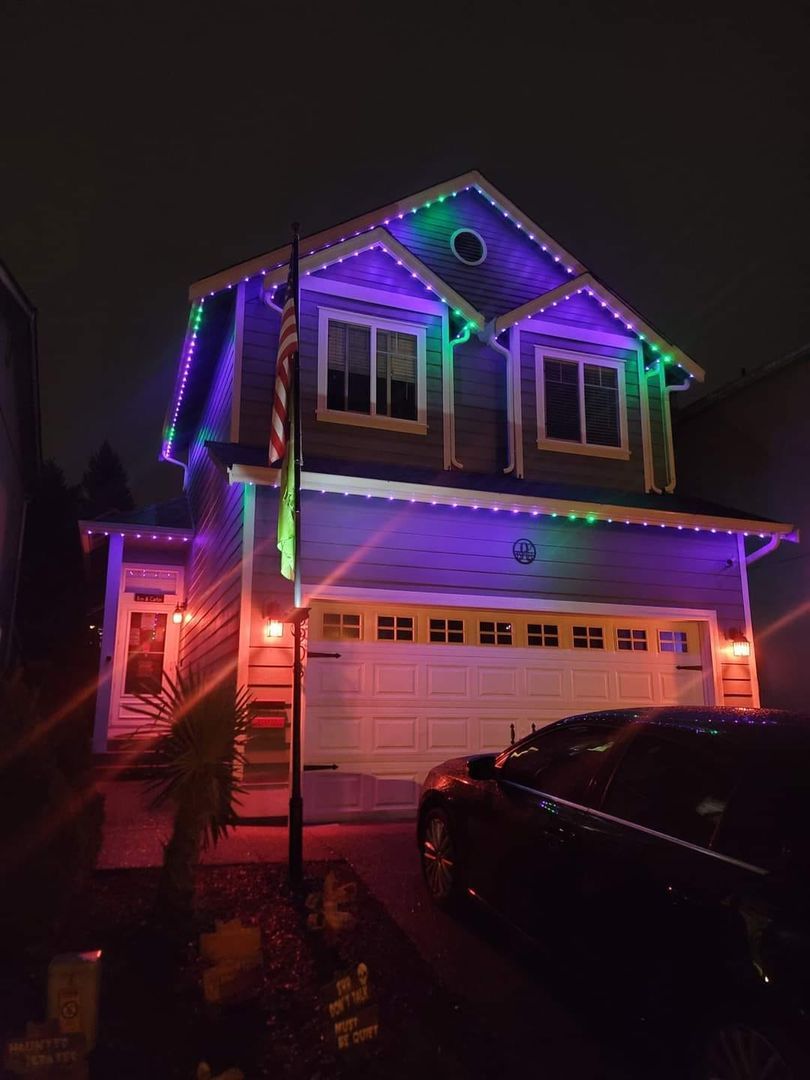 A two-story house at night lit by permanent multicolored holiday lights with an American flag and a parked car in the driveway