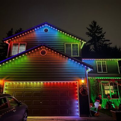 A two-story house at night with multi-colored lights along the roof and an inflatable Santa decoration near the window.