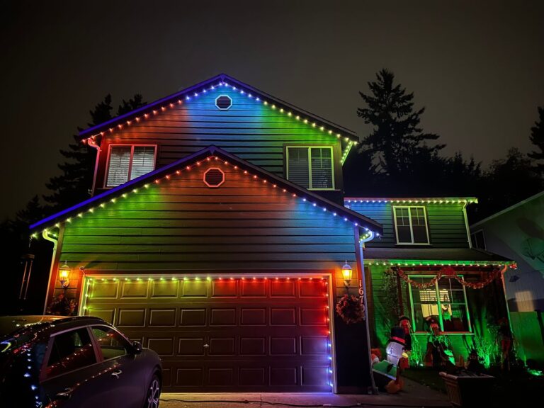 A two-story house at night with multi-colored lights along the roof and an inflatable Santa decoration near the window.