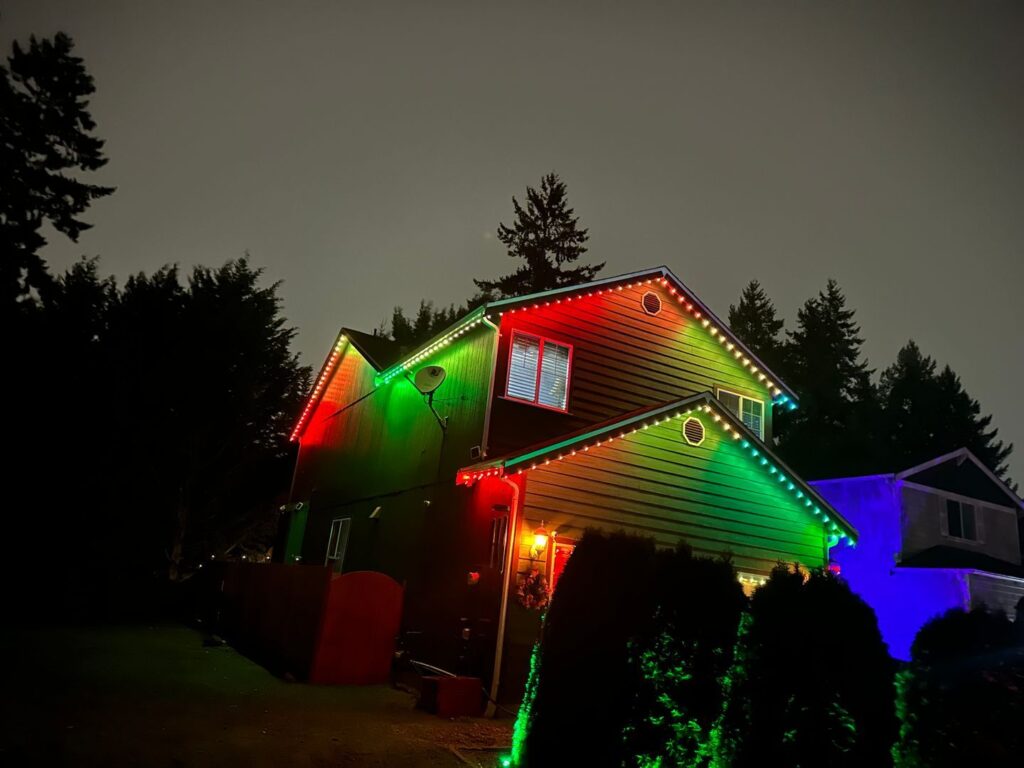 A two-story house at night illuminated by red, green, and blue lights along its eaves against a backdrop of trees.