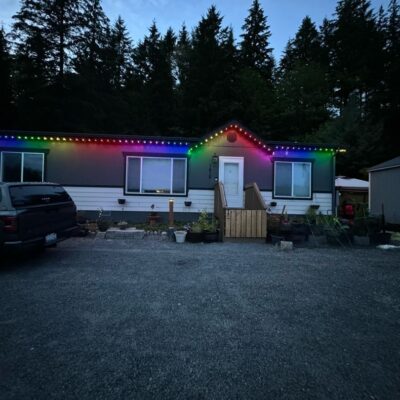 Single-story house with permanent multi-colored lights along the roofline, gravel driveway, parked truck, and surrounding trees.