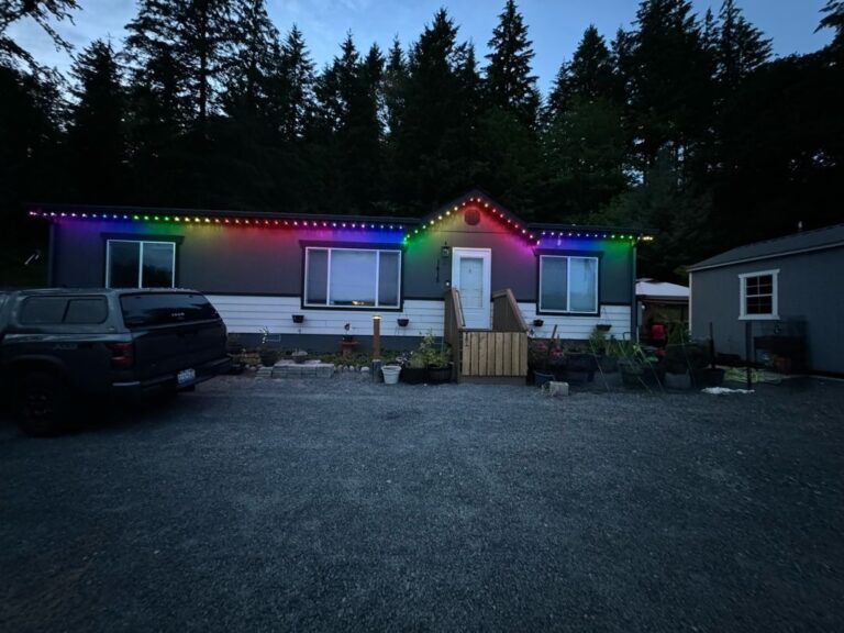 Single-story house with permanent multi-colored lights along the roofline, gravel driveway, parked truck, and surrounding trees.