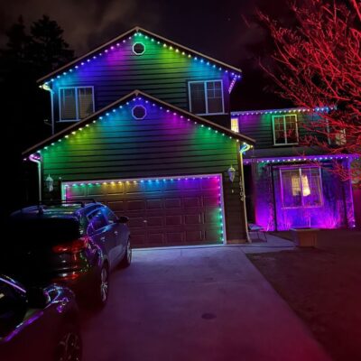 Two-story house decorated with multi-colored string lights at night, with two cars parked in the driveway.