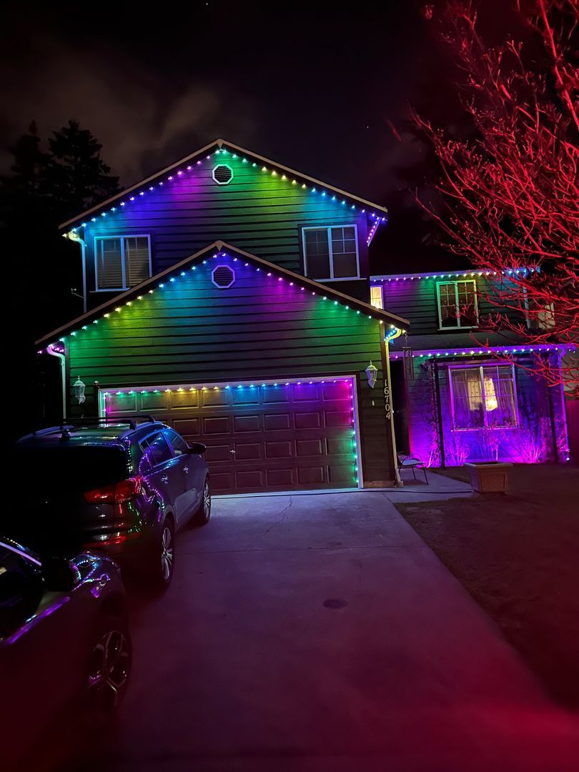 Two-story house decorated with multi-colored string lights at night, with two cars parked in the driveway.