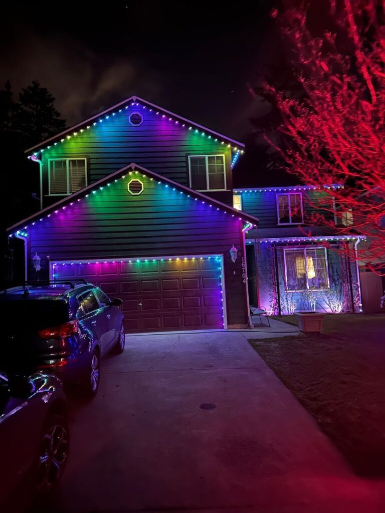 Two-story house with multicolored LED lights and cars in the driveway at night.