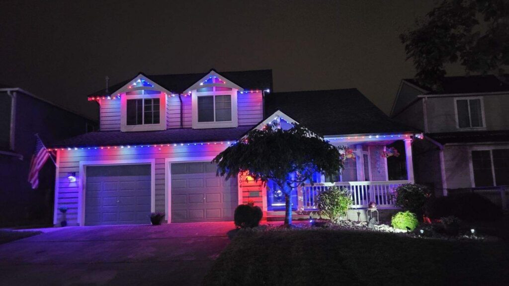 Two-story suburban house decorated with red, blue, and white lights, featuring an American flag and illuminated tree.