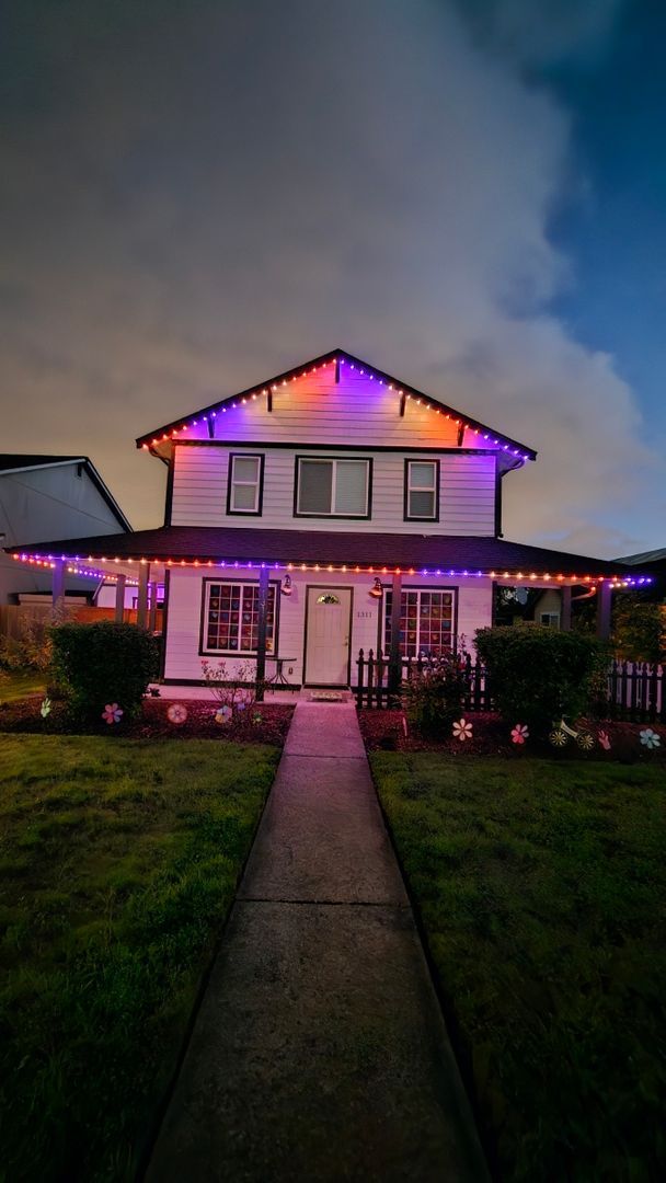 Two-story house with multicolored holiday lights on the roofline, white exterior, black trim, picket fence, and flower decorations in the garden.